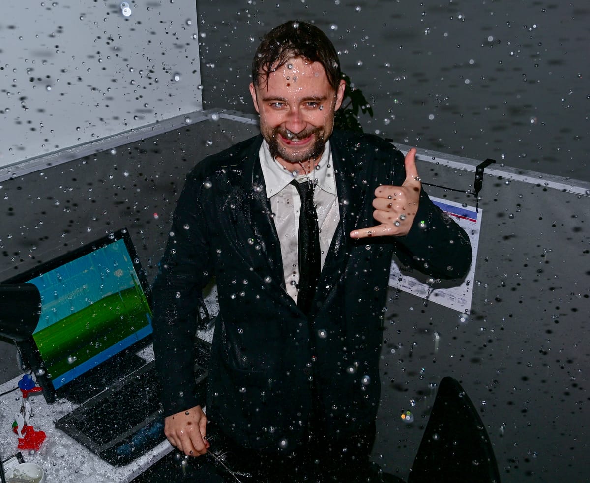 Lukas Bentel, soaked, throwing a shaka at a desk in the rain